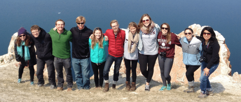 A group of students pose together on a rocky cliff overlooking a large lake dotted with ice.
