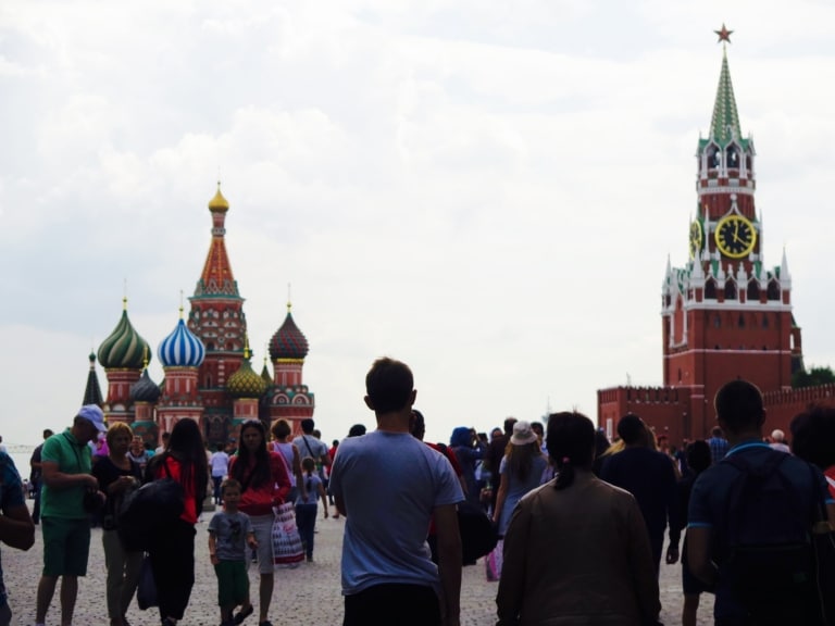Crowds of tourists walking through Red Square in Moscow, with St. Basil's Cathedral and a Kremlin tower in the background.