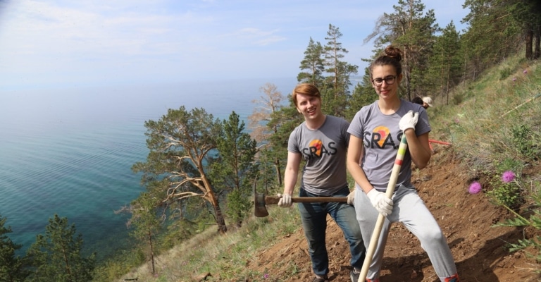 Two students in SRAS t-shirts holding tools on a hillside above Lake Baikal.