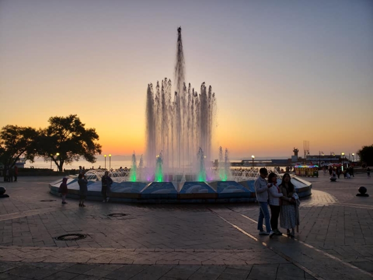 A colorfully lit fountain in a plaza at dusk, with people gathered nearby and the sea visible in the background.
