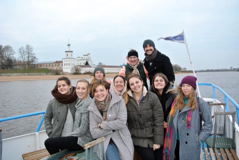 A group of students and their program leader posing on a riverboat, with a white monastery visible on the shore behind them.