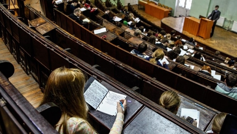 Students take notes in a large university lecture hall as a lecturer speaks from the podium below.