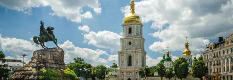 The Bohdan Khmelnytsky Monument and St. Sophia's bell tower in Kyiv, with golden domes visible under a partly cloudy sky.
