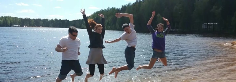 A group of students jump joyfully into the air at a lake shore surrounded by forest.