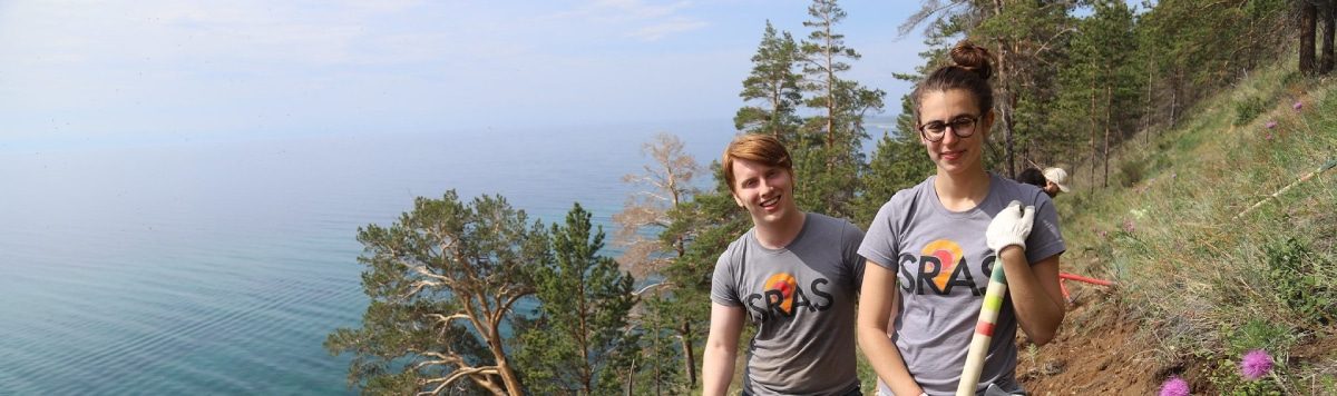 Two students wearing SRAS t-shirts do trail maintenance work on a forested hillside above Lake Baikal.