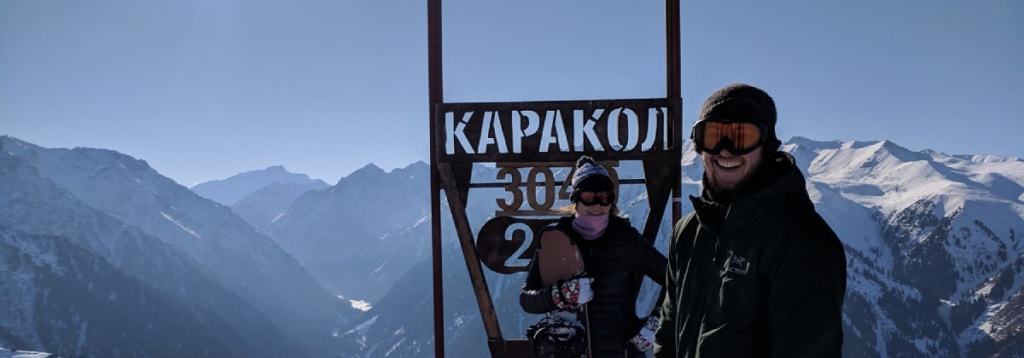 Two snowboarders pose at the Karakol ski resort sign in the snow-capped mountains of Kyrgyzstan.