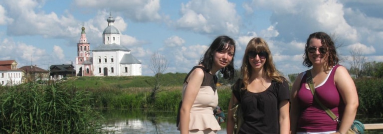 Three students stand by a pond with a historic white church visible across the water in Suzdal, Russia.