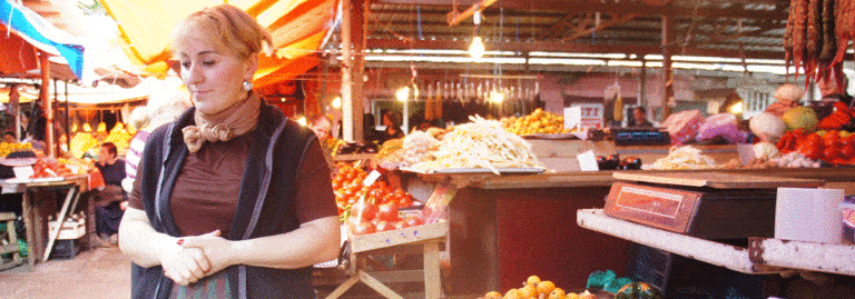 A woman stands amid produce and food stalls inside a bustling open-air market.