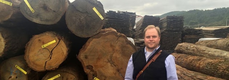 Simone standing in front of stacked logs at a timber yard.