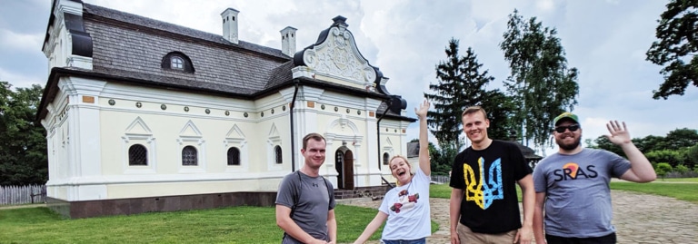 Four students posing and waving in front of a historic white palace building in Ukraine.