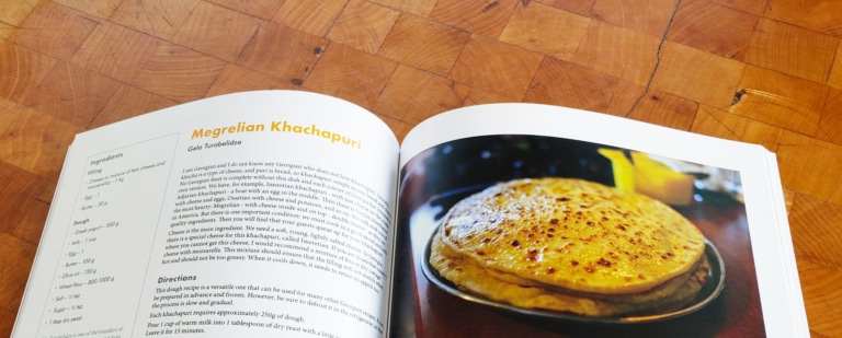 Open pages of the SRAS commemorative cookbook displaying a khachapuri recipe on a wooden table.