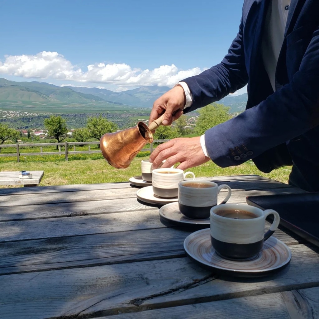 A man pouring Armenian coffee from a copper cezve into small cups at an outdoor table overlooking mountains.