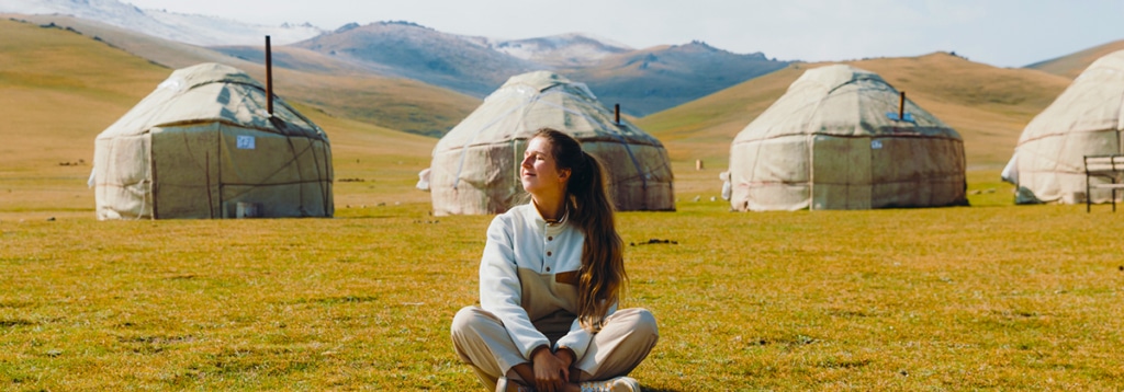 A peacefully smiling woman sitting cross-legged in front of a row of yurts in a grassy valley.