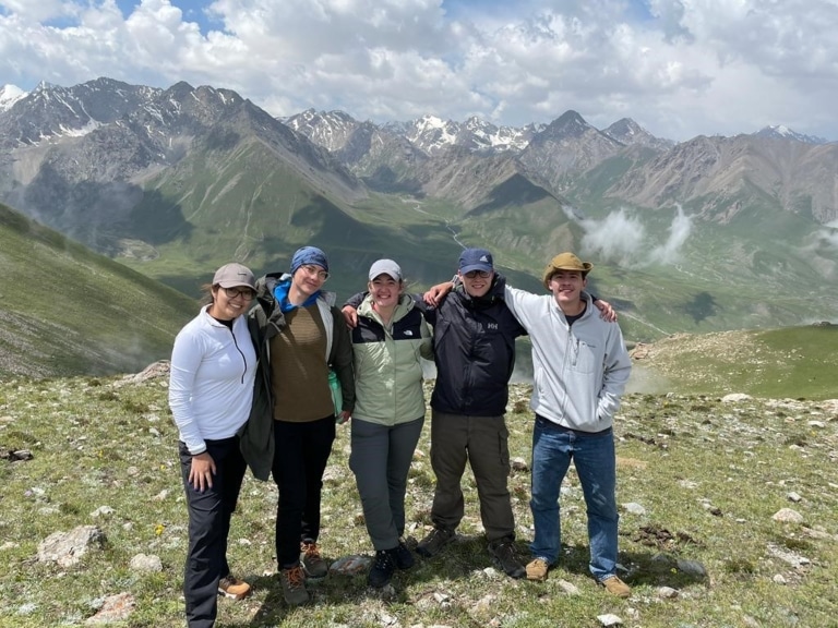 Five students posing together on a grassy ridge overlooking a mountain valley with wisps of mist in Kyrgyzstan.