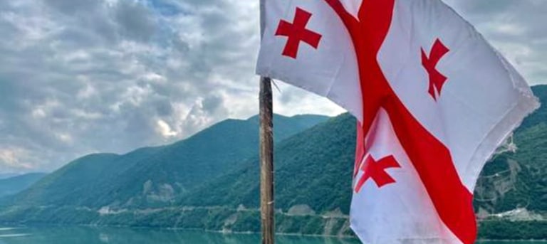 The Georgian flag waving in front of mountains and a lake at the Jinvali Reservoir.
