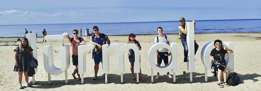A group of people posing in front of large letters spelling "Jurmala" on a beach in Latvia.