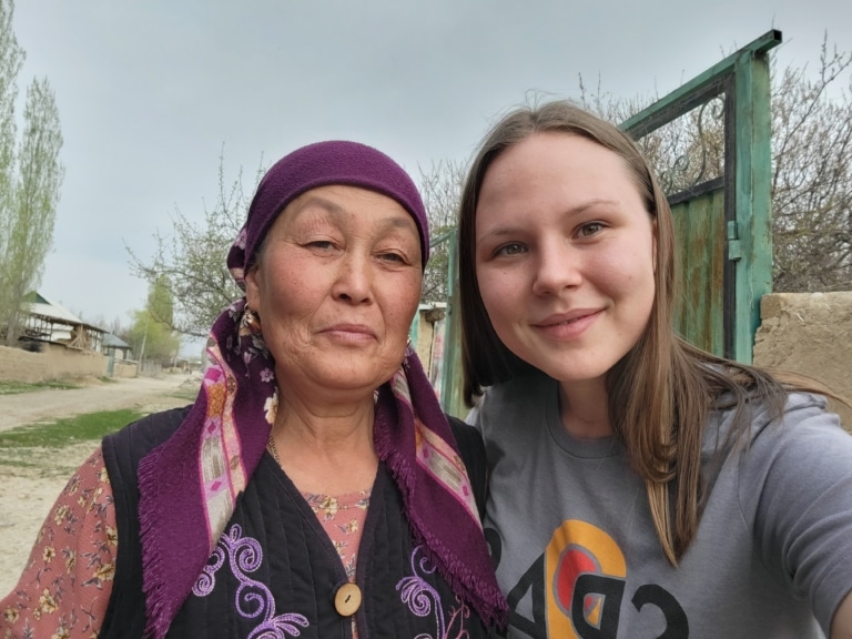 A student and an elderly woman in a purple headscarf taking a selfie together outdoors in a village.