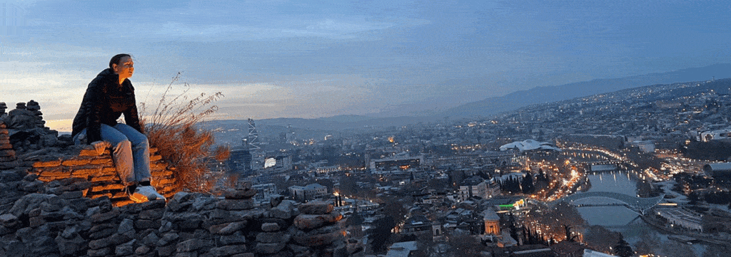 A woman sitting on ancient stone ruins overlooking Tbilisi's illuminated skyline at dusk.