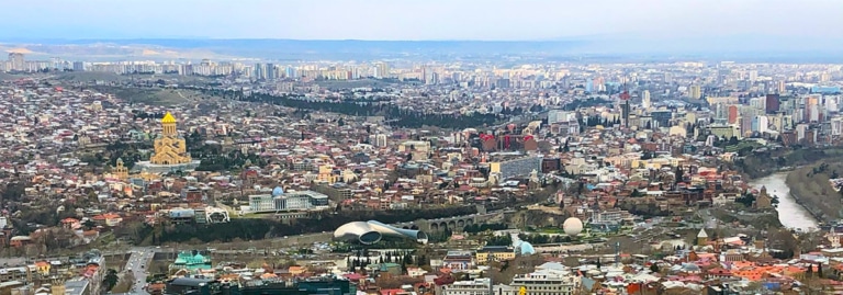 An aerial view of Tbilisi's skyline, with the golden-domed Sameba Cathedral visible on a hill.