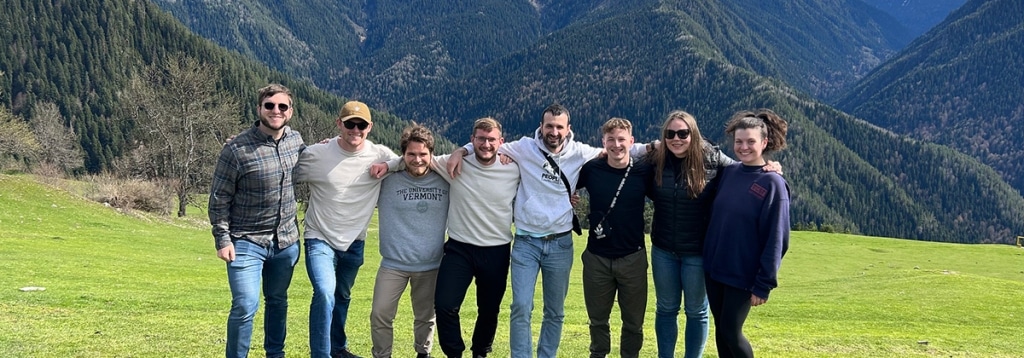 Eight students pose arm-in-arm together in front of green mountains during a study abroad trip.