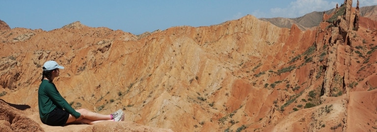 A woman sitting on a rock overlooking the red rock formations of Skazka Canyon in Kyrgyzstan.