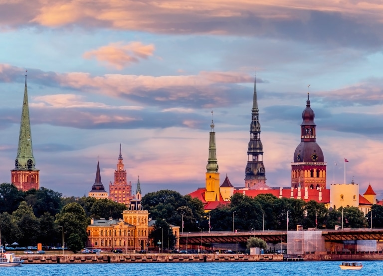 The spires of Riga's Old Town skyline reflected in warm sunset light, viewed from across the river.