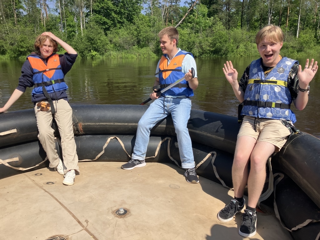 Three teenagers wearing life jackets posing on an inflatable raft on a calm river in Latvia.