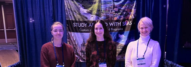 Three smiling people standing in front of 'Study Abroad with SRAS' banner'.