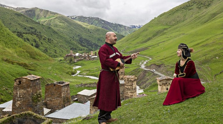 panduri lute while a woman in traditional attire looks on near the historic stone towers of Ushguli, Georgia, in the Caucasus Mountains.