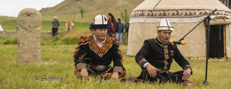 kalpak hats and embroidered velvet garments, seated near a traditional felt yurt and an ancient stone balbal (balbal is a carved stone statue from nomadic Turkic cultures) during a cultural event or nomad festival in Kyrgyzstan.