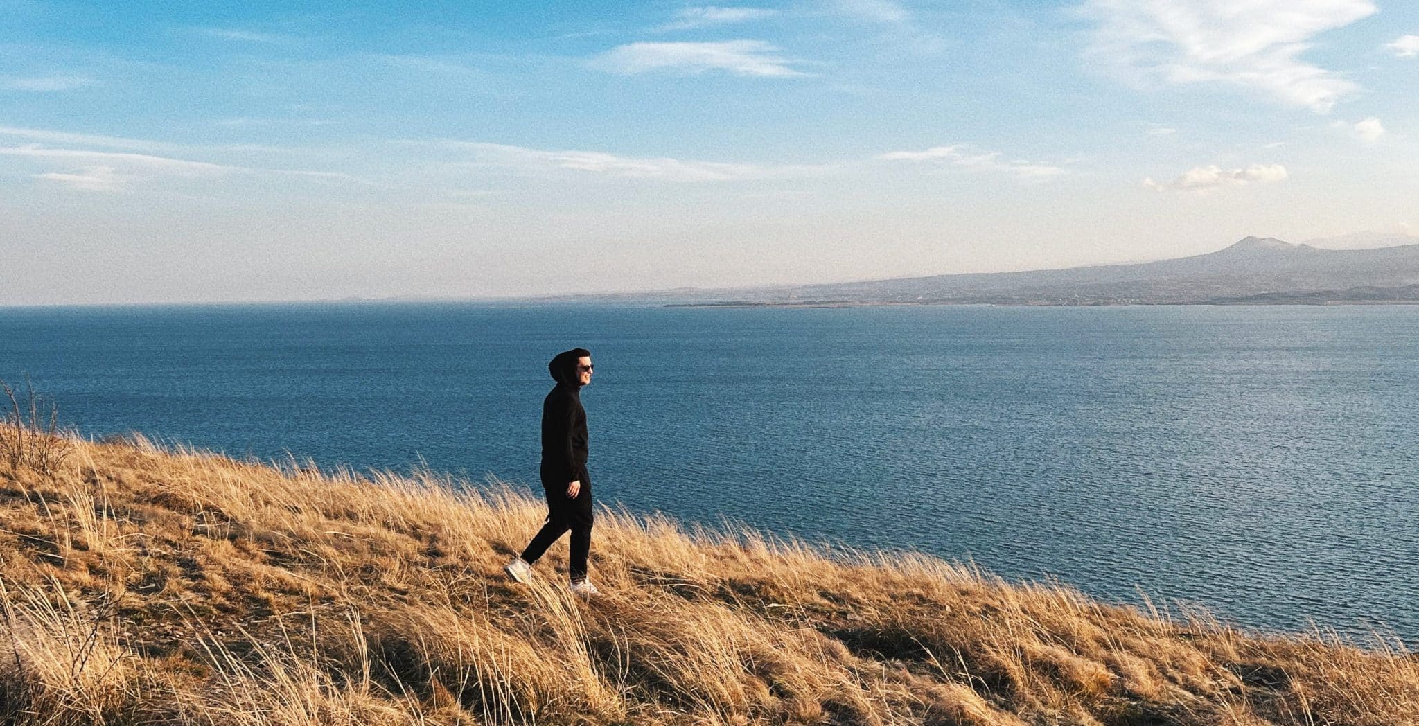 Man walking in yellow grass by deep blue lake.