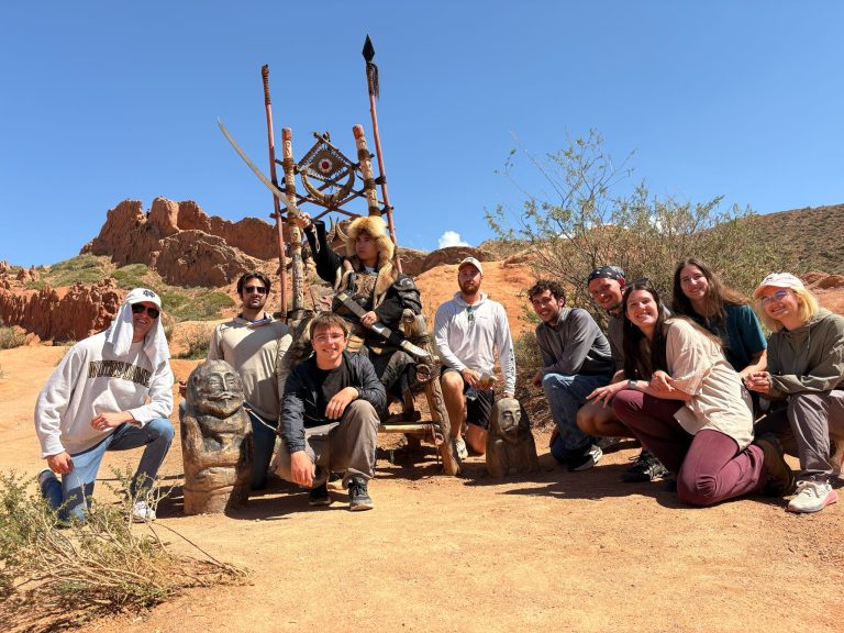 Students posing with man in traditional dress in desert landscape.