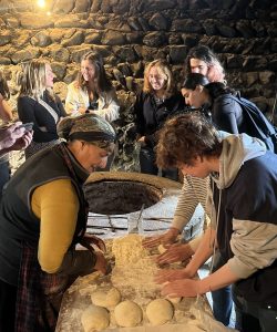 Students being taught to roll dough in stone-walled room by woman in traditional dress.
