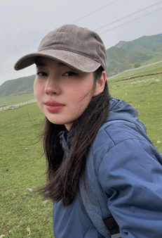 Casually dressed young woman with Asian features in green field with mountains in background.