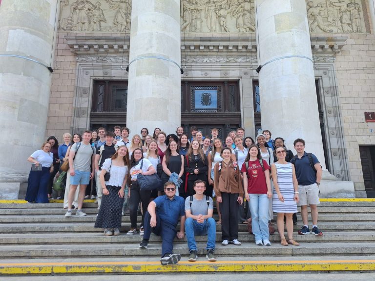 Group of smiling students in front of historic stone building with columns and carved relief.