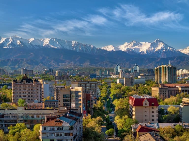 View of colorful buildings amidst trees with snowcapped mountains in background on a sunny day.