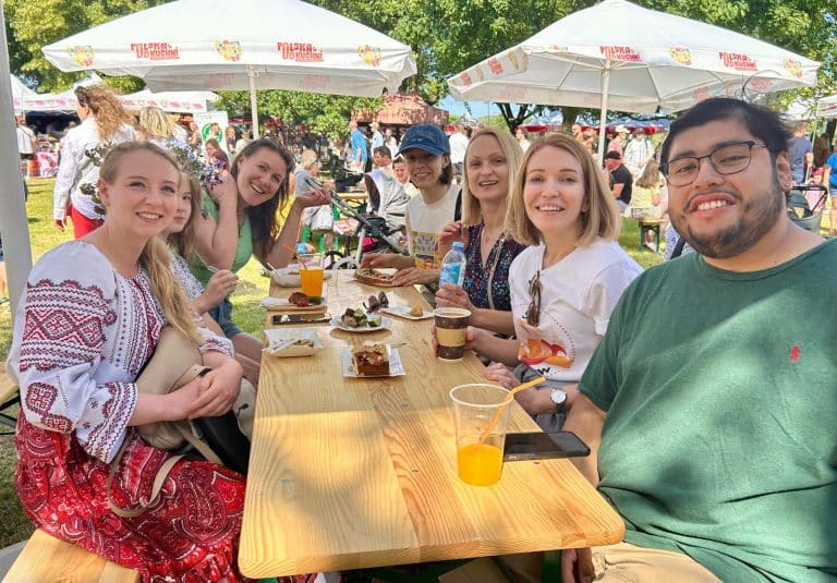 a diverse group of people sitting around a long wooden outdoor table at a daytime Ukrainian food festival in Warsaw, with white market umbrellas overhead and various snacks, pastries, and drinks on the table.