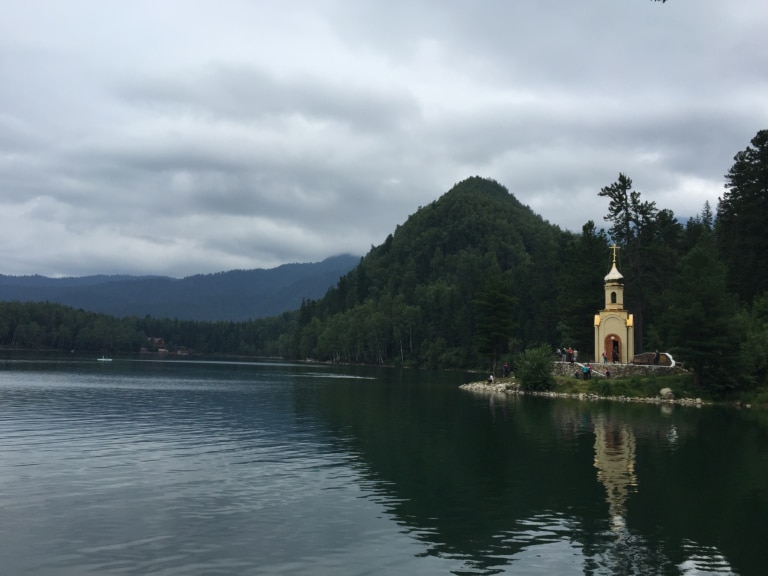 A lakeside chapel near Lake Baikal in Irkutsk, Russia, surrounded by forested hills and visitors.